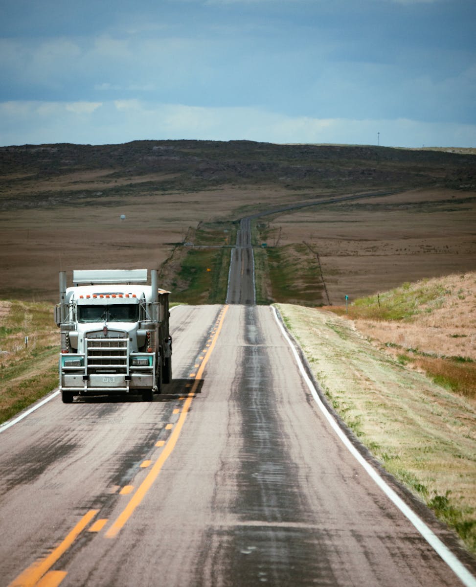 A truck traveling on a long empty highway through expansive countryside under a blue sky.